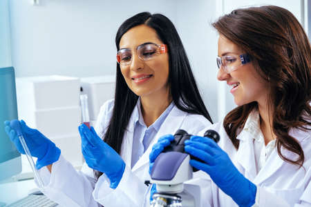 Two beautiful young female scientists working in laboratory with test tubes and microscope doing...