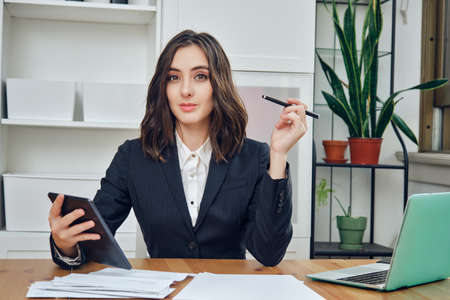 thoughtful beautiful young adult businesswoman manager in a suit with a tablet pc in officeの写真素材
