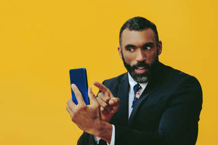 portrait of angry handsome bearded man in suit and tie with smartphone video call pointing finger at...