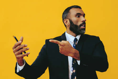 portrait of angry expressive excited bearded man in suit and tie with smartphone video call pointing...