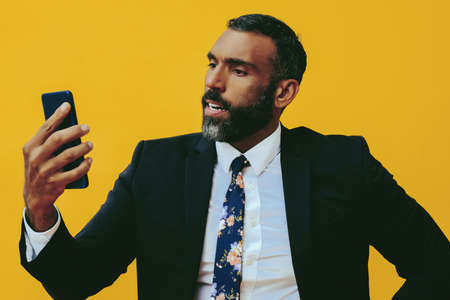 portrait of angry expressive bearded man in suit and tie with smartphone video call yellow backgroundの写真素材