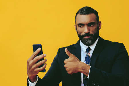 portrait of smiling positive handsome bearded man in suit and tie with smartphone video call thumbs up on yellow backgroundの写真素材