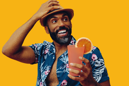 happy bearded mid adult african american man wearing Hawaiian shirt and hat smiling at orange juice cocktail looking up at copy space on yellow backgroundの写真素材