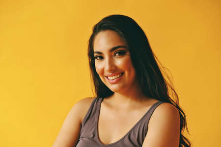 headshot beauty shot of beautiful hispanic Latina woman smiling young adult with black long hair and tank top in front of yellow background looking at cameraの写真素材