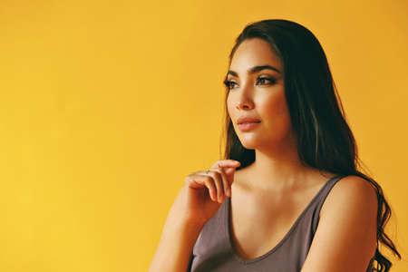 headshot beauty shot of thoughtful hispanic Latina woman young adult with black long hair and tank top hand on chin in front of yellow background looking away at copy spaceの写真素材