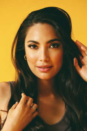 headshot beauty shot of sensual hispanic Latina woman with young adult with hands on black long hair and tank top in front of yellow background looking at cameraの写真素材