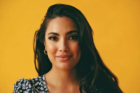 headshot of sensual thoughtful hispanic Latina woman young adult with black long hair and dress in front of yellow background looking at cameraの写真素材