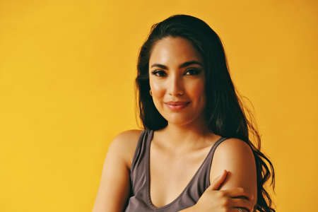 headshot beauty shot of sensual hispanic Latina woman smiling young adult with black long hair and tank top in front of yellow background looking at cameraの写真素材