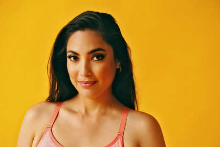 headshot beauty shot of hispanic Latina woman smiling smart young adult with black long hair and tank top in front of yellow background looking at cameraの写真素材