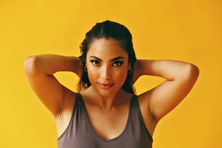 headshot beauty shot of hispanic Latina woman smiling young adult with hands on hair black long hair and tank top in front of yellow background looking at cameraの写真素材