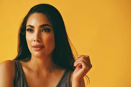headshot beauty shot of hispanic Latina woman smiling young adult with black long hair and tank top in front of yellow background looking away at copy spaceの写真素材