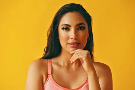 headshot beauty shot of thoughtful hispanic Latina woman hand on chin young adult with black long hair and tank top in front of yellow background looking at cameraの写真素材