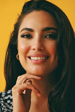 headshot beauty shot of sensual smiling hispanic Latina woman with hand on chin young adult with black long hair and dress in front of yellow background looking at cameraの写真素材