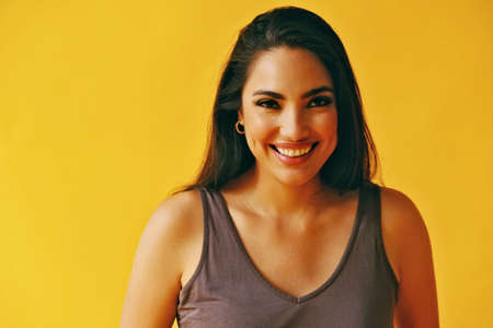 headshot beauty shot of hispanic Latina woman toothy smile young adult with black long hair and tank top in front of yellow background looking at cameraの写真素材