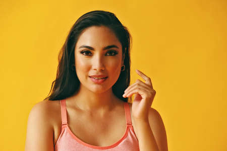 headshot beauty shot of thoughtful hispanic Latina woman smiling young adult with black long hair and tank top in front of yellow background looking at cameraの写真素材