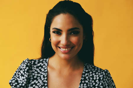 headshot beauty shot of sensual smiling hispanic Latina woman young adult with black long hair and dress in front of yellow background looking at cameraの写真素材