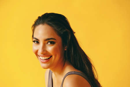 headshot beauty shot of hispanic Latina woman toothy smile young adult with black long hair and tank top in front of yellow background looking at cameraの写真素材