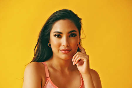 headshot beauty shot of thoughtful hispanic Latina woman hand on chin young adult with black long hair and tank top in front of yellow background looking at cameraの写真素材