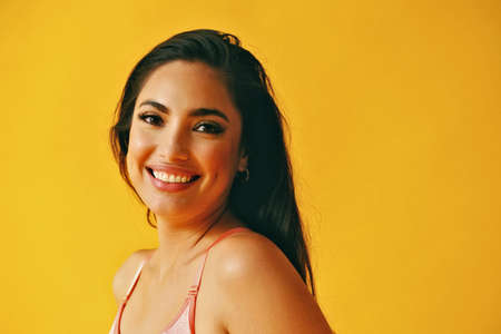 headshot beauty shot of hispanic Latina woman toothy smile young adult with black long hair and tank top in front of yellow background looking at cameraの写真素材