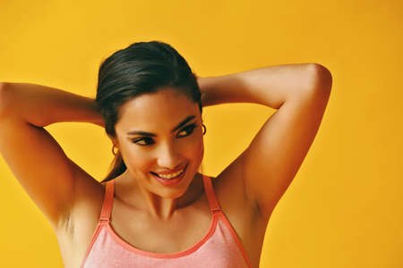 headshot beauty shot of hispanic Latina woman smiling young adult with hands on hair black long hair and tank top in front of yellow background looking away at copy spaceの写真素材