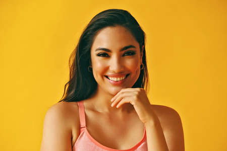 headshot beauty shot of hispanic Latina woman with toothy smile hand on chin young adult with black long hair and tank top in front of yellow background looking at cameraの写真素材