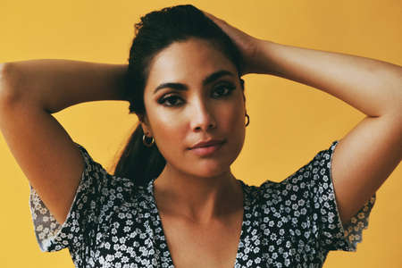 headshot beauty shot of sensual thoughtful hispanic Latina woman young adult with hands on black long hair and dress in front of yellow background looking at cameraの写真素材