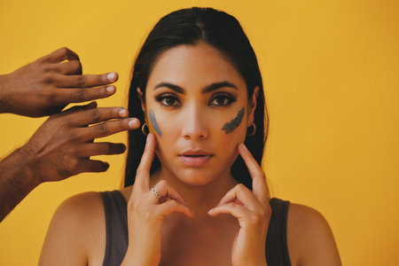 Beauty shot portrait of hispanic Latina woman applying clay mud with fingers and hands around face young adult black long hair and tank top in front of a yellow background looking at cameraの写真素材