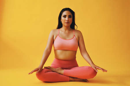 waist up portrait of hispanic beauty Latina woman hands on knees sports outfit young adult in front of yellow background looking at cameraの写真素材