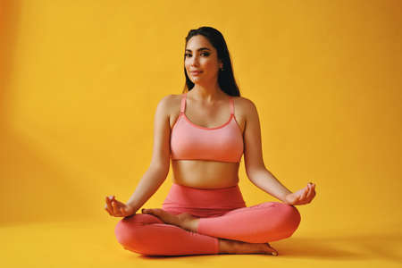 portrait of hispanic beauty Latina woman sitting cross legged in yoga pose sports outfit young adult in front of yellow background looking away at copy spaceの写真素材