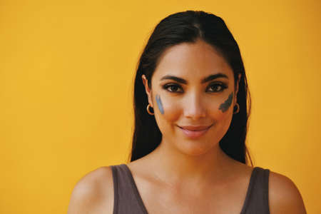 Close up Beauty shot portrait of hispanic Latina woman with clay mud mask on cheek smiling young adult black long hair and tank top in front of a yellow background looking at cameraの写真素材