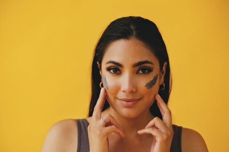 Beauty shot portrait of hispanic Latina woman applying clay mud with finger young adult black long hair and tank top in front of a yellow background looking at cameraの写真素材