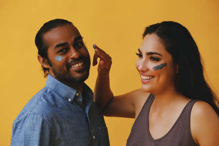 portrait of mid adult playful couple hispanic Latina woman and Indian Man applying clay mud with fingers and hands laughing smiling in front of a yellow background looking at cameraの写真素材