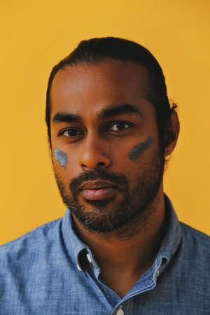 Close up Beauty shot portrait of thoughtful Indian Man with clay mud mask on cheek young adult black hair and blue shirt in front of a yellow background looking at cameraの写真素材