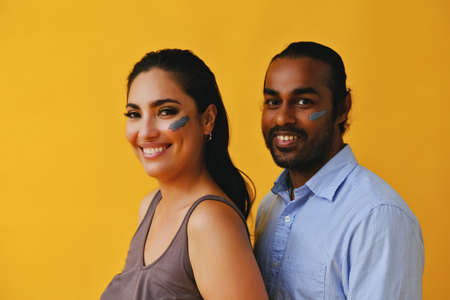 portrait of mid adult playful couple hispanic Latina woman and Indian Man with clay mud on cheeks and hands laughing smiling in front of a yellow background looking at cameraの写真素材
