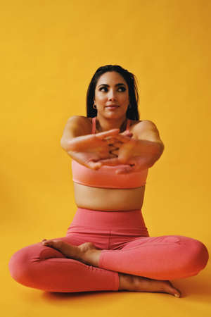 portrait of hispanic beauty Latina woman sitting cross legged in yoga pose sports outfit stretching arms young adult in front of yellow background looking away at copy spaceの写真素材