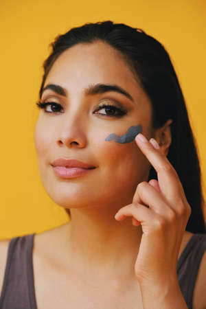 Close up Beauty shot portrait of hispanic Latina woman applying clay mud with finger young adult black long hair and tank top in front of a yellow background looking at cameraの写真素材