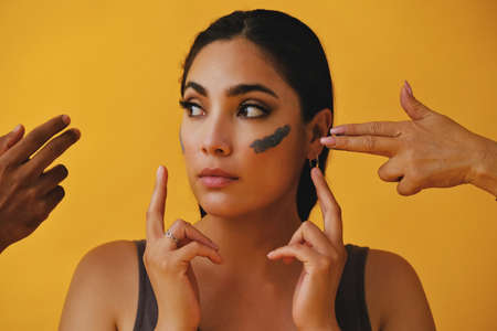 Beauty shot portrait of hispanic Latina woman applying clay mud with fingers and hands around face young adult black long hair and tank top in front of a yellow background looking away at copy spaceの写真素材