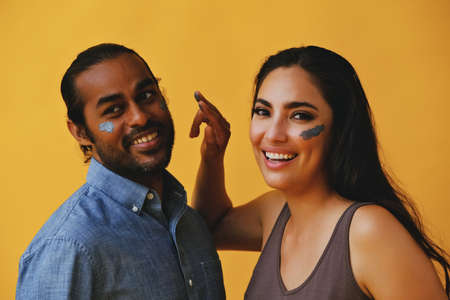 portrait of mid adult playful couple hispanic Latina woman and Indian Man applying clay mud with fingers and hands laughing smiling in front of a yellow background looking at cameraの写真素材