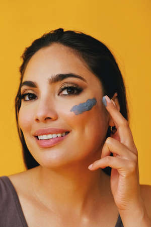 Close up Beauty shot portrait of hispanic Latina woman applying clay mud with finger young adult black long hair and tank top in front of a yellow background looking at cameraの写真素材