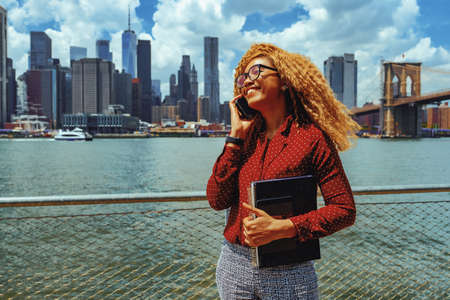 Portrait young adult entrepreneur millennial woman with eyeglasses and afro hair talking on a phone call outdoors with Manhattan New York City skyline behindの写真素材