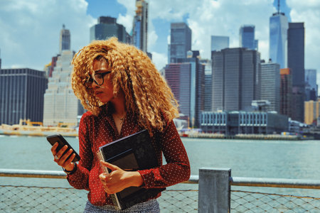 Portrait young adult entrepreneur millennial woman with eyeglasses and afro hair in a video call conference thoughtful outdoors with Manhattan New York City skyline behindの写真素材