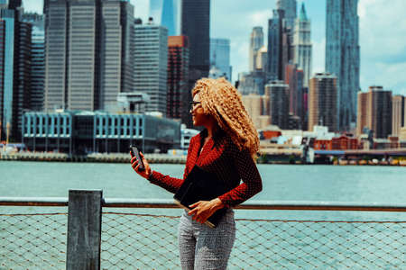 Portrait young adult entrepreneur millennial woman with eyeglasses and afro hair in a video call conference outdoors with Manhattan New York City skyline behindの写真素材
