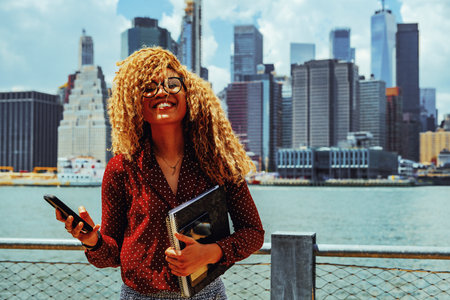 Portrait young adult entrepreneur millennial woman with eyeglasses and afro hair smiling at camera outdoors with Manhattan New York City skyline behindの写真素材