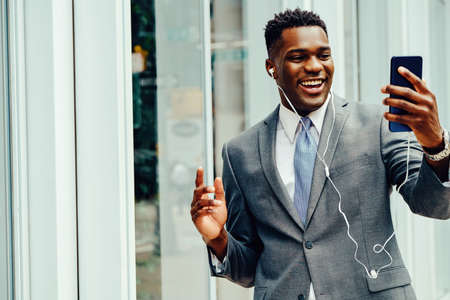 Positive young businessman waving hand at smartphone outside wearing suitの写真素材