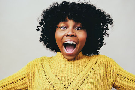 Closeup portrait of cheerful Black woman laughing open mouth looking at camera with open arms wearing yellow sweater isolated against gray backgroundの写真素材