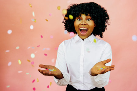 Confetti throw- Black woman with big smile celebrates success and happiness. Studio shot over pink backgroundの写真素材