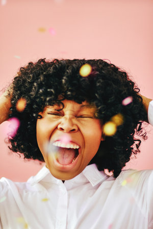 Happiness closeup portrait of cheerful beautiful black young adult woman wearing white shirt with open mouth and hands on hair under confetti rain celebrating isolated over pink backgroundの写真素材