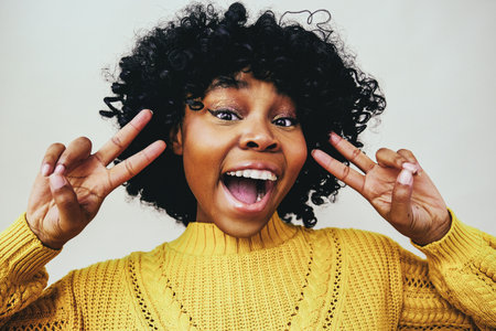 closeup portrait of a happy black woman smiling with open mouth and fingers in victory sign next to face wearing a yellow sweater against a gray backgroundの写真素材