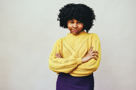 Good or bad decisions. Black woman with arms crossed looking at camera wearing a yellow sweater isolated against a gray backgroundの写真素材