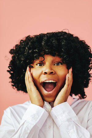 Close-up portrait of cheerful young adult black woman excited with open mouth and hands on cheeks isolated on pink backgroundの写真素材